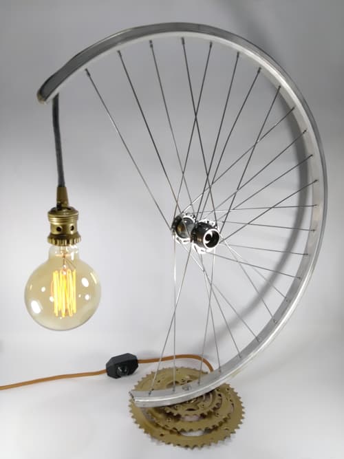 Pieza decorativa de iluminación realizada con llanta y plato de bicicletas recuperados. Cable textil color ocre, interruptor con dimer y portalámparas pintado de color oro, al igual que la base. 